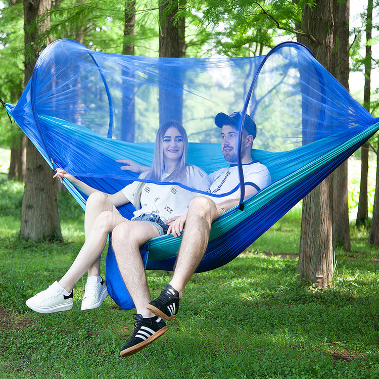 Two people sitting in a blue hammock with mosquito net in a forest setting